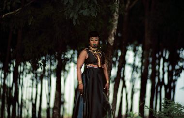 Djely Tapa portant robe noir, marron, bleu et motifs multicolores, colliers africains, maquillage de scène sur fond extérieur d'arbres