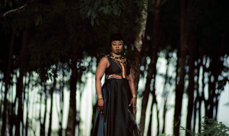 Djely Tapa portant robe noir, marron, bleu et motifs multicolores, colliers africains, maquillage de scène sur fond extérieur d'arbres