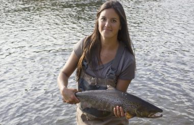 A woman stands next to a body of water holding a salmon.