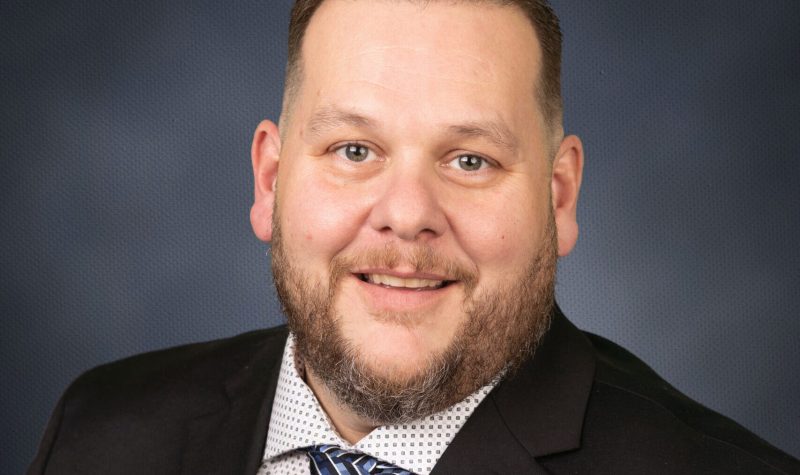 A man with short dark hair, trimmed beard and mustache and wearing a dark suit with a white shirt and blue tie is shown against a dark blue background.