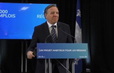 Quebec Premier Francois Legault in a dark suit behind a blue podium and in front of a Quebec flag and blue background.