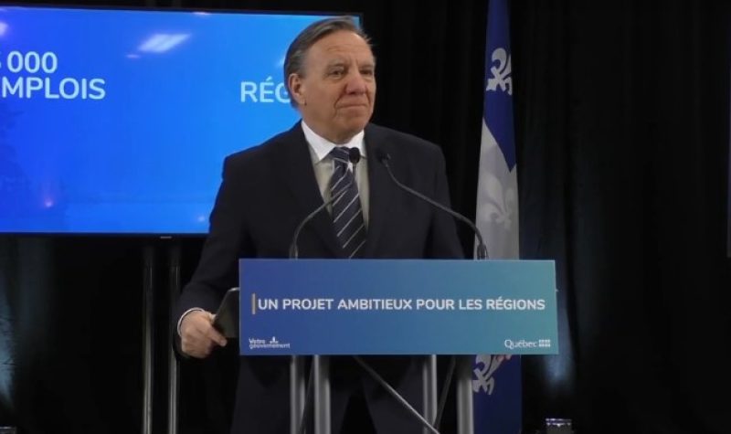 Quebec Premier Francois Legault in a dark suit behind a blue podium and in front of a Quebec flag and blue background.