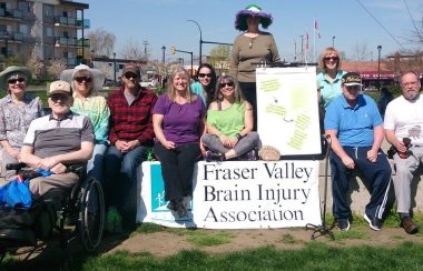 A group of people sit outside with signs on a sunny day and pose for a picture for the Fraser Valley Brain Injury Association.