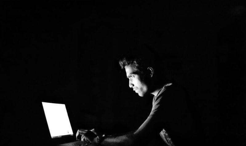 Un homme de côté et dans la pénombre, uniquement éclairé par un écran d'ordinateur portable, tape quelque chose sur le clavier de celui-ci.
