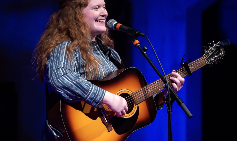 A young woman with long red hair holding a guitar stands at a microphone.