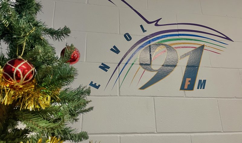 Le logo de la radio communautaire Envol91 FM en couleur bleu marine, avec des lignes en forme d'oiseau aux couleurs violet, bleu, vert, or et rose. À côté du logo se trouve un arbre de Noël auquel sont accrochées des décorations.