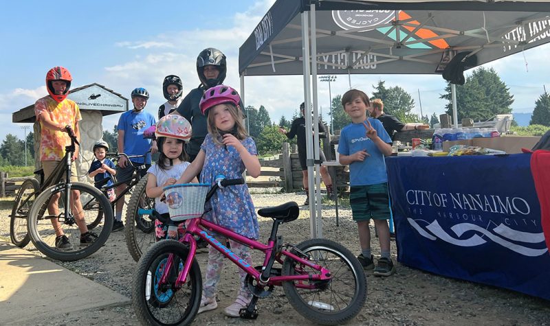 A group of children with bicycles at a station with a tent and a city of Nanaimo banner draped over a table.