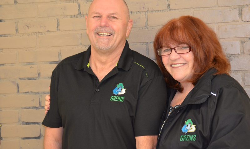 A man and woman wearing black shirts with a blue, green and white GREINS logo on them stand in front of a beige brick wall.