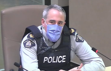 A man wearing a COVID face mask, police vest and uniform, sitting next to a microphone.