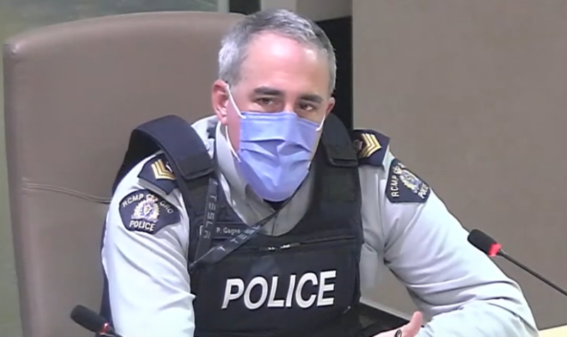 A man wearing a COVID face mask, police vest and uniform, sitting next to a microphone.