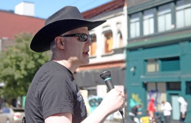 Garth Mullins stands at a memorial on a sunny street holding a microphone and wearing a black cowboy hat