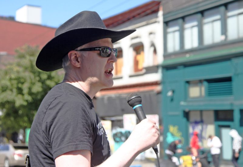 Garth Mullins stands at a memorial on a sunny street holding a microphone and wearing a black cowboy hat