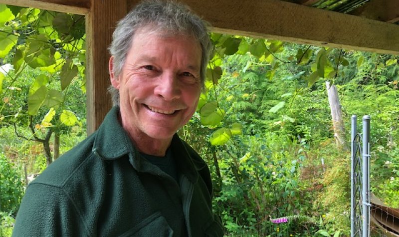 A smiling man with grey hair wearing a green shirt stands on a deck with forest in the background.