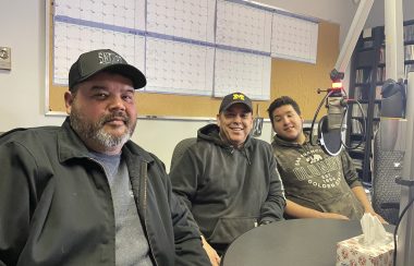 Three men sit in sound studio around a microphone at CKRZ FM