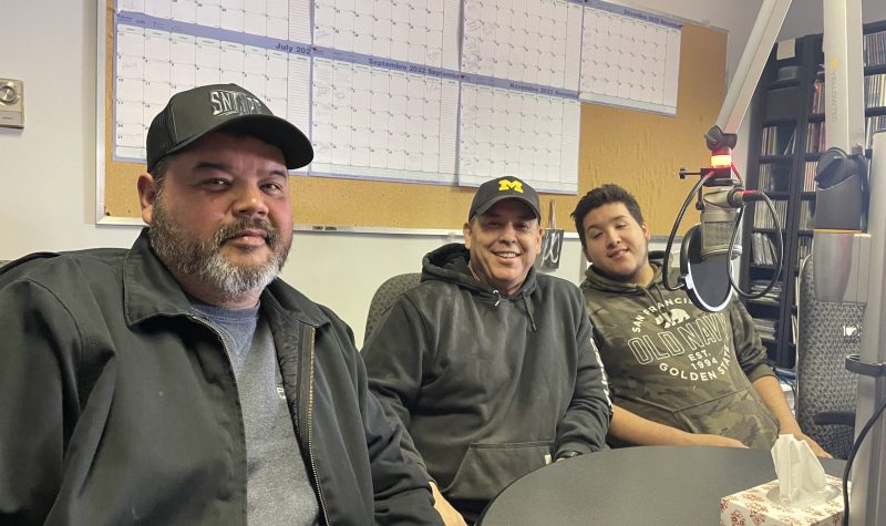 Three men sit in sound studio around a microphone at CKRZ FM