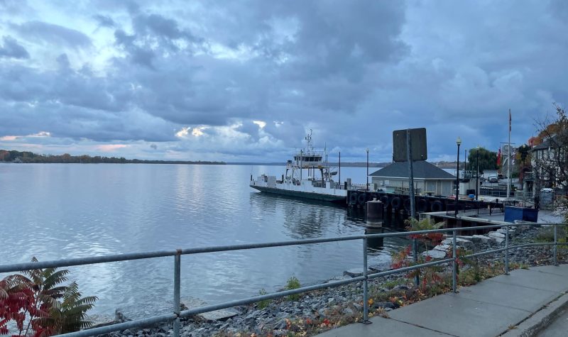 A landscape perspective photo of a lakefront with sidewalk, metal railing, fallen autumnal leaves, and shoreline protective rocks in the immediate foreground. Beyond is the lake and to the right is a small white painted vehicle ferry that is disembarking from its dock. Buildings and infrastructure cluster around the dock.