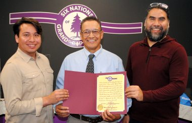 Three men pose for a picture. The man in the middle wearing a blue collared shirt and black tie holds a folder showing the document for the camera. In the background a purple and white logo for Six Nations of the Grand River is shown featuring a purple tree.