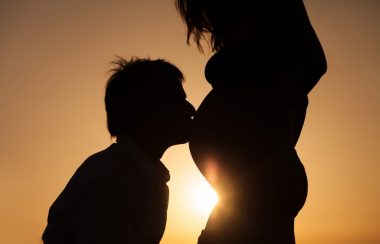 À contre-jour devant un coucher de soleil, un homme donne un baiser sur le ventre d'une femme enceinte.