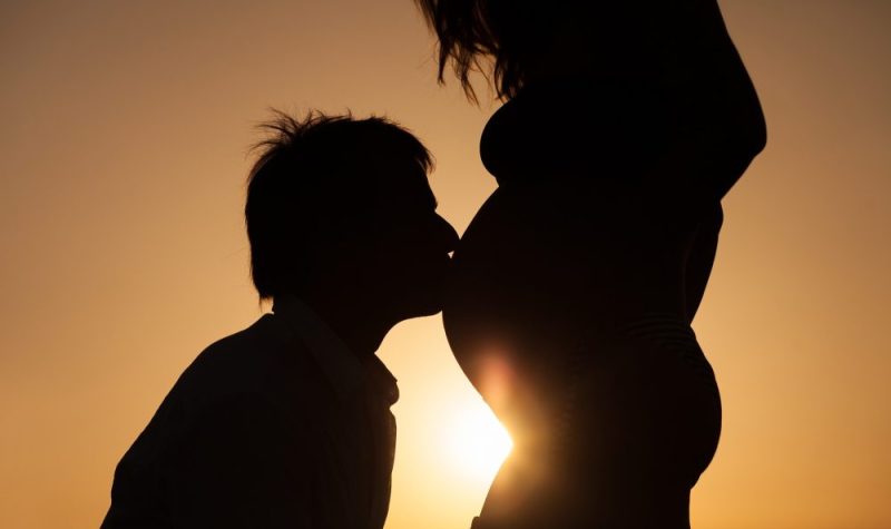 À contre-jour devant un coucher de soleil, un homme donne un baiser sur le ventre d'une femme enceinte.