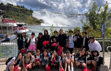On peut voir le groupe des 18 étudiantes japonaises photographiées devant les chutes du Niagara. Les étudiantes sont souriantes et on peut voir les chutes à l'arrière ainsi que le bateau servant à faire les excursions vers les chutes.