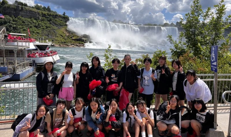 On peut voir le groupe des 18 étudiantes japonaises photographiées devant les chutes du Niagara. Les étudiantes sont souriantes et on peut voir les chutes à l'arrière ainsi que le bateau servant à faire les excursions vers les chutes.