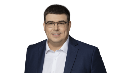 Pictured is a headshot of Paquet. He is standing against a white background wearing a black suit, white dress shirt, and rectangular glasses.