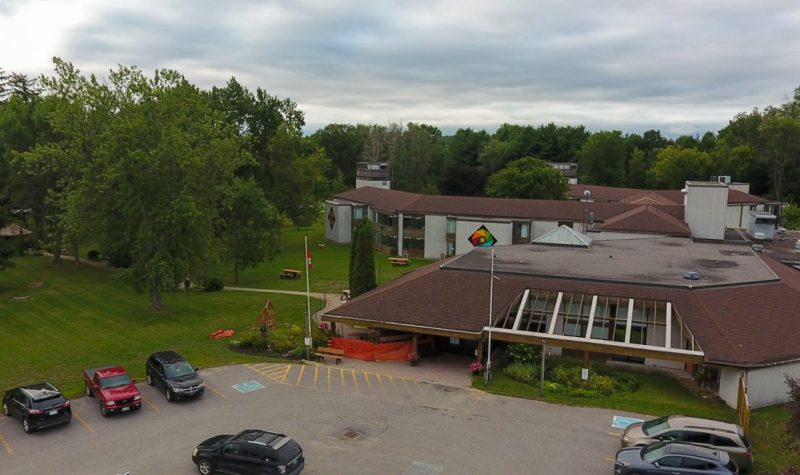 A picture of HJ McFarland long-term care facility. It depicts a grey bricked building with a low setting brown roof on the right side. To the left is a green space with a lawn and trees. In the lower foreground is a parking lot with cars parked in rows.