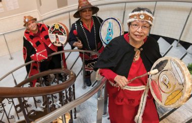 Three Indigenous people walk up a spiral staircase beating traditional drums