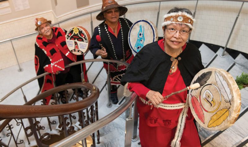 Three Indigenous people walk up a spiral staircase beating traditional drums