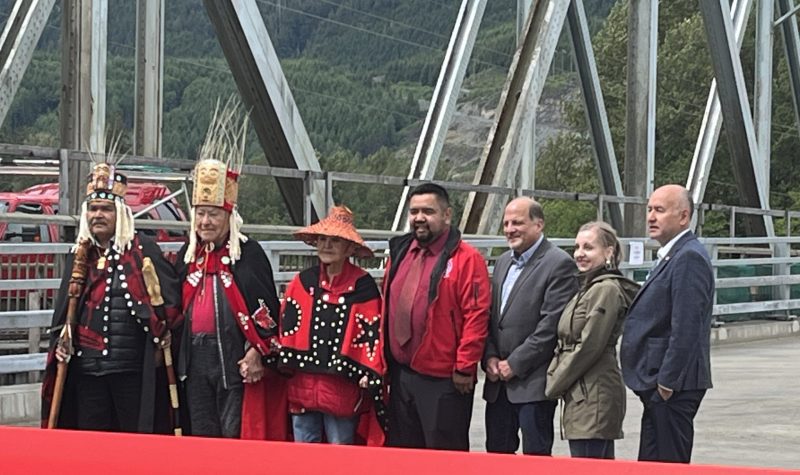 A row of people including the hereditary chiefs and dignitaries along with political officials on the new Haisla Bridge. They are standing behind a large red ribbon.