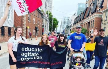 The Halifax Workers' Action Centre (WAC) are a non-profit seen walking the streets with a banner that says educate and agitate, there are about 30 people in the background. It is during the day during the summer.