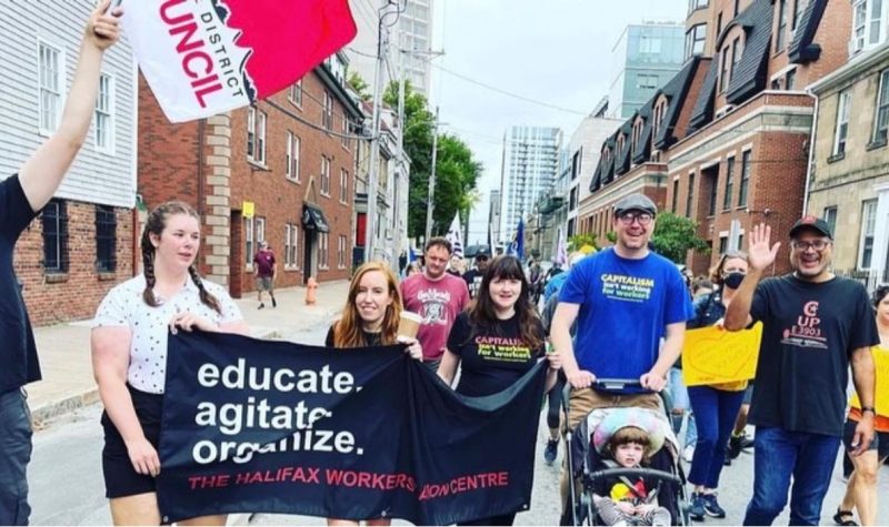 The Halifax Workers' Action Centre (WAC) are a non-profit seen walking the streets with a banner that says educate and agitate, there are about 30 people in the background. It is during the day during the summer.