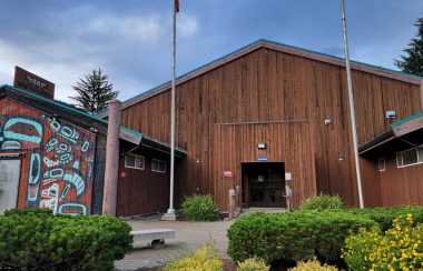 A wooden building with two smaller attachments in the front, the left on has an indigenous design on the front.