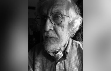 A black-and-white photo shows an elderly man with white hair and a white beard wearing glasses.