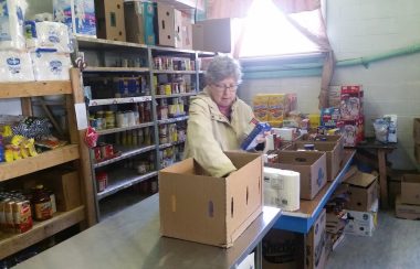 A woman unpacks a box of food supplies, with shelves of food in the background.