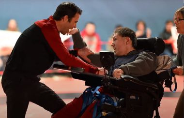 A boy in a wheelchair pushed by a woman confronts a man dressed in a red sweater as part of a professional wrestling match.