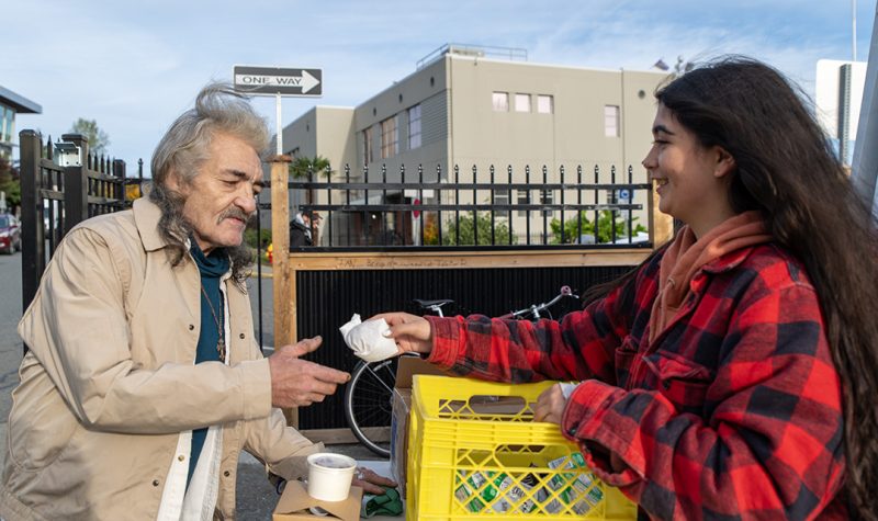 A man in a beige jacked is handed a wrapped piece of food from a woman wearing a red plaid jacket.