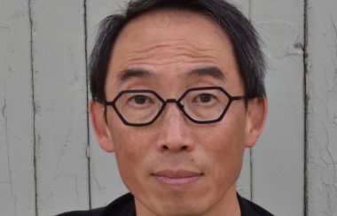 A portrait of artist Henry Tsang in a black shirt and against a white, wood panel background.