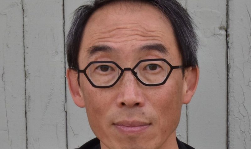 A portrait of artist Henry Tsang in a black shirt and against a white, wood panel background.