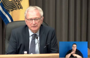 A man sitting at a desk with a New Brunswick flag behind him, a inset of a person doing sign language.