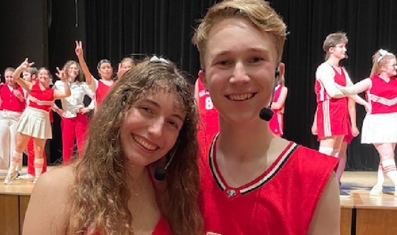 A young woman with long brown a young man with short blonde hair stand side-by-side smiling. Behind them a number of people dressed in red and white athletic clothes stand on a stage, some of them gesturing to the camera. There is a dark curtain behind them.