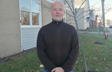 A man stands outside a building and in front of a tree. He is wearing a brown sweater and denim jeans and has his hands folded in front of him.