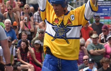 Homme habillé d'un chandail de hockey jaune, blanc et bleu avec un casque de hockey bleu ayant les bras levé devant une foule.