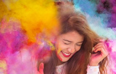 A woman is seen standing in a cloud of colourful powder being thrown into the air. She is smiling, with her eyes closed.