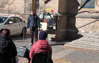 A person speaking into a microphone in front a brick building as people sitting down watch them.