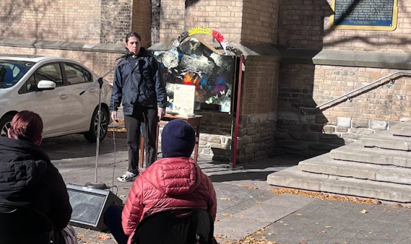 A person speaking into a microphone in front a brick building as people sitting down watch them.