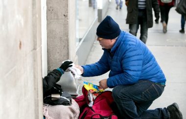 A man wearing a blue jacket, black pants and a black toque hands what appears to be a towel to a homeless person