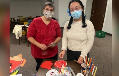 Two women wearing face masks are shown in a church basement, standing at a table working on crafts.