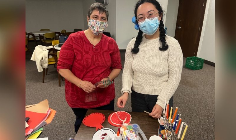 Two women wearing face masks are shown in a church basement, standing at a table working on crafts.