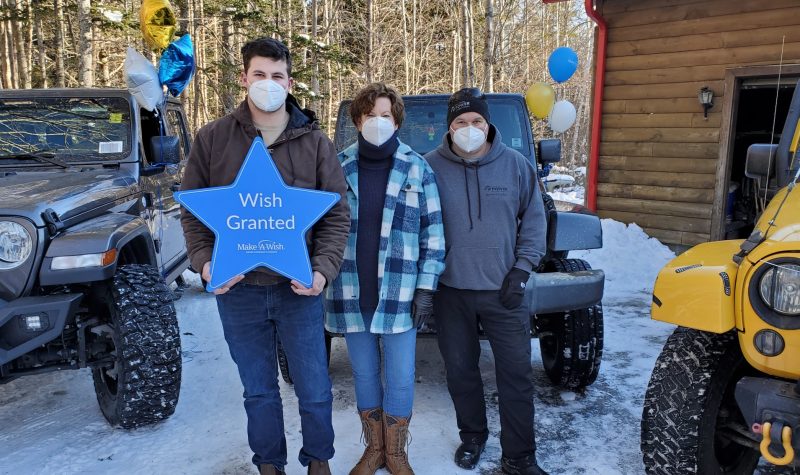 Three people stand in front of a jeep. One holds a blue Make-A-Wish star in his hands.